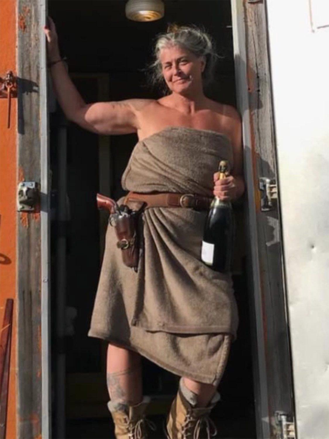 Sue Aikens After the World's Best Camp Shower – Kavik River Camp Alaska Sue Aikens stepping out of the Kavik River Camp bathhouse wrapped in a towel, wearing a gun holster and holding a bottle of champagne, smiling in the Arctic summer light